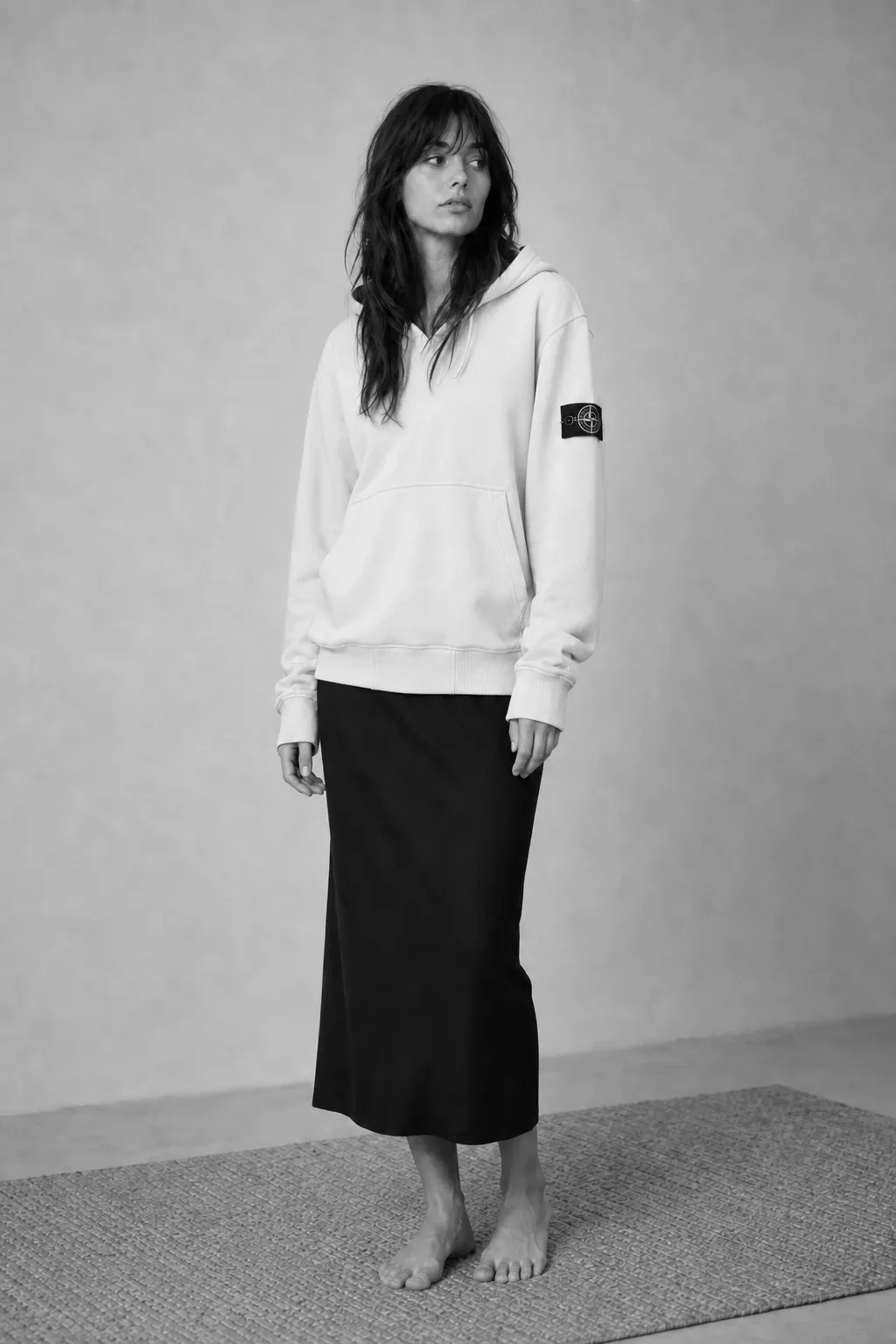 Black-and-white portrait of a woman wearing a light Stone Island pullover hoodie with a dark midi skirt, standing barefoot in a studio.