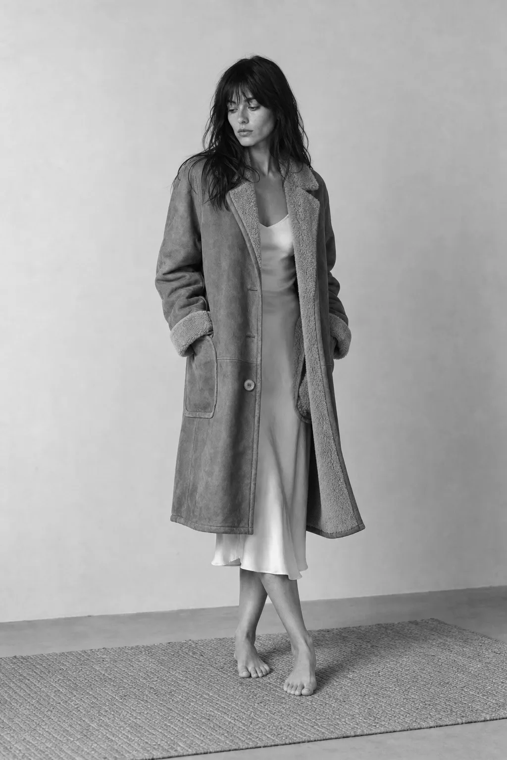 A woman with long dark hair stands barefoot on a narrow rug in a pale studio, wearing an open knee-length shearling | The Weight of Winter, Relover